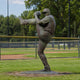 Life-Size Contemporary Baseball Pitcher Bronze Sculpture by Giant Sculptures, 240cm figure mid wind-up with raised knee, displayed on grass beside a ball field.