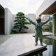 Monumental Contemporary Astronaut Bronze Sculpture by Giant Sculptures with arms outstretched and verdigris patina, displayed beside a reflecting pool and bonsai in a concrete courtyard.