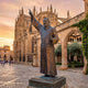 Giant Sculptures Large Traditional Robed Figure Bronze Sculpture 210cm, arm raised skyward and holding a cross, displayed on a plinth outside a Gothic cathedral.