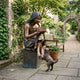 Life-Size Traditional Girl on Bench with Dog Bronze Sculpture by Giant Sculptures displayed on a stone patio beside a wooden bench and potted geraniums.