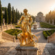 Gold-polished bronze Monumental Classical Standing Male Figure and Leopard Sculpture by Giant Sculptures, displayed on a plinth in a formal garden with fountain.