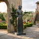 Life-Size Traditional Girl with Bird Trays Bronze Sculpture by Giant Sculptures standing in a Tuscan courtyard with roses, stone archway, and steps.