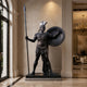 Life-Size Classical Greek Warrior Bronze Statue by Giant Sculptures displayed in a marble lobby, holding a tall spear and round shield with plumed helmet.