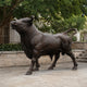 Life-Size Traditional Bull Bronze Sculpture by Giant Sculptures shown outdoors on stone paving, head raised and tail curled, with trees and a building behind.