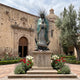 Giant Sculptures Monumental Our Lady of Guadalupe Bronze Statue in polychrome teal and cream with spike halo, on a stone pedestal in a rose-filled church courtyard.