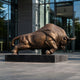 Monumental Charging Bull Bronze Sculpture by Giant Sculptures, 320cm, shown in charging pose on a dark granite plinth outside a modern glass building.