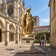 Monumental Classical Virgin Mary Bronze Statue by Giant Sculptures in polished gold finish on a stone pedestal in a Gothic cathedral courtyard with visitors nearby.