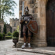 Monumental Heraldic Armoured Knight Bronze Sculpture by Giant Sculptures, 320cm, holding a raised sword and U-emblem shield beside a castle archway.