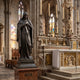 Monumental Classical Virgin Mary Bronze Statue by Giant Sculptures, 300cm, hands crossed at heart with dark patina, displayed beside a Gothic cathedral altar.