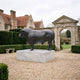 Life-Size Traditional Standing Bull Bronze Sculpture with dark patina on a white marble plinth, displayed on gravel beside a stone archway and manor house.