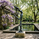 Life-Size Nude Ribbon Dancer Bronze Sculpture, 240cm, in deep verdigris patina on a stone plinth beside a garden pool with wisteria and trees.