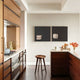 A modern kitchen features wooden cabinetry with white countertops, a sleek stool near the island, and a gray wall decorated with Modulus Black & White Duo Panel Abstract Wall Art by Giant Sculptures. A bowl of oranges, a cup, and bright flowers in a vase complete the scene.