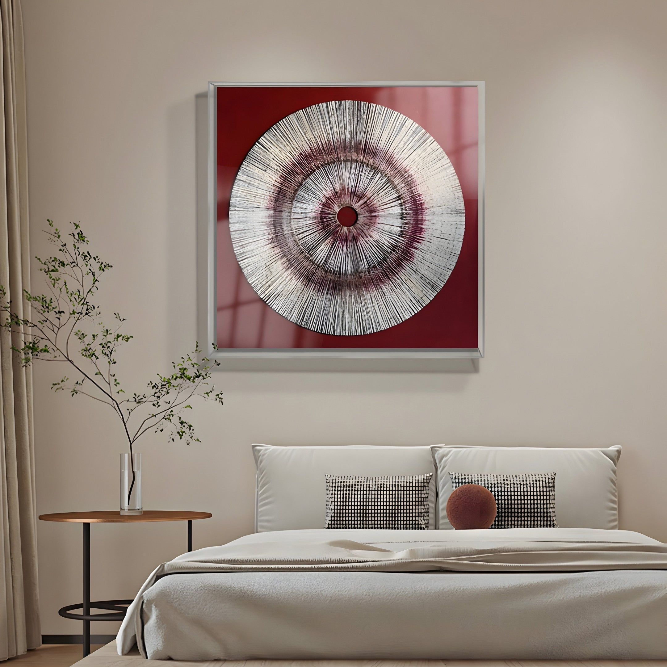 A minimalist bedroom includes Giant Sculptures Oculis Crimson Aura Square Handmade 3D Paper Wall Art with a circular design on red. Beneath the art is a bed adorned with white and patterned pillows, adjacent to a small round wooden table with a slender vase and branches.