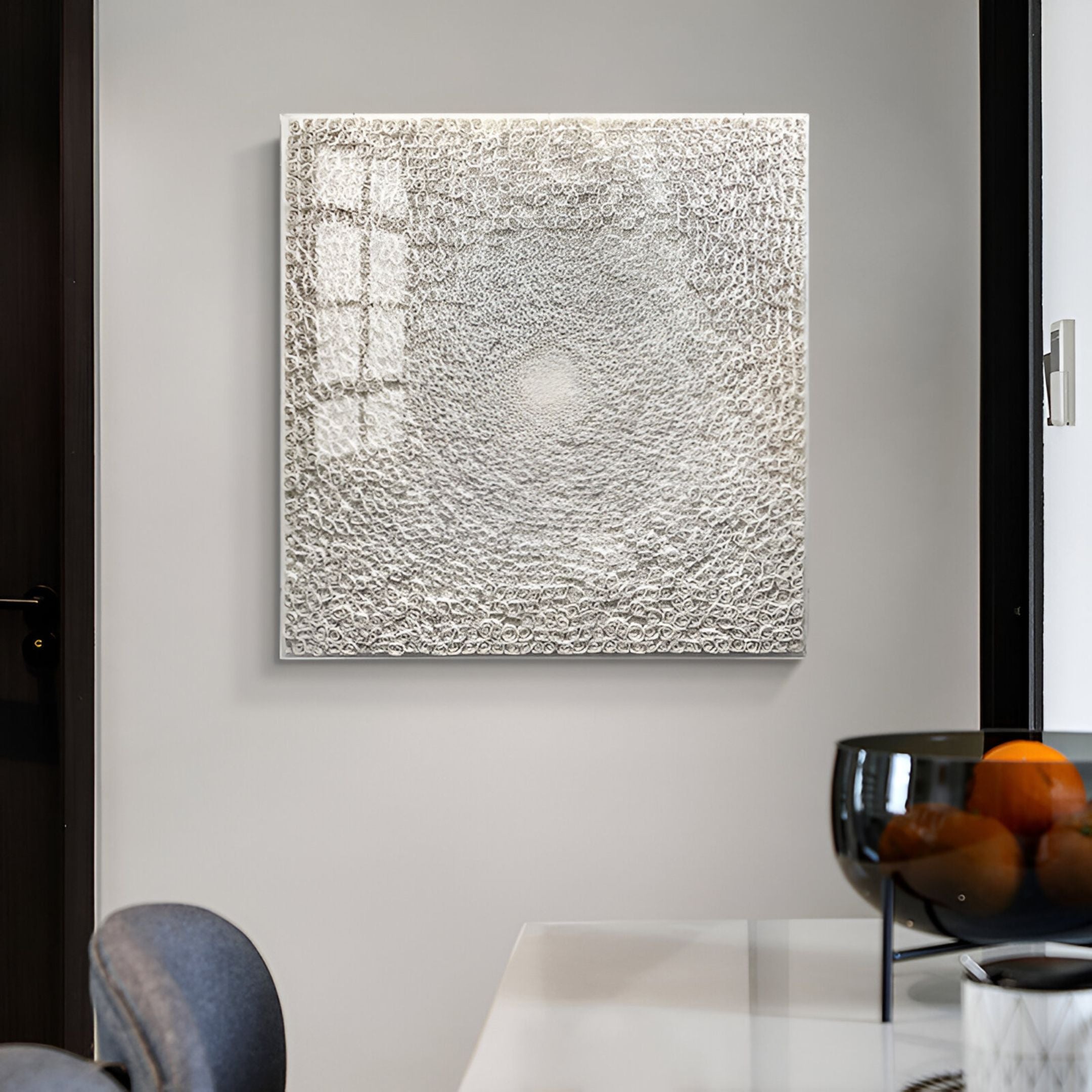 Mounted on a light gray wall, the TerraLuma Ivory White Bloom Paper Craft 3D Wall Art by Giant Sculptures displays intricate swirling patterns in a textured circular design. In the foreground sits a modern dining table adorned with a dark glass bowl of oranges.