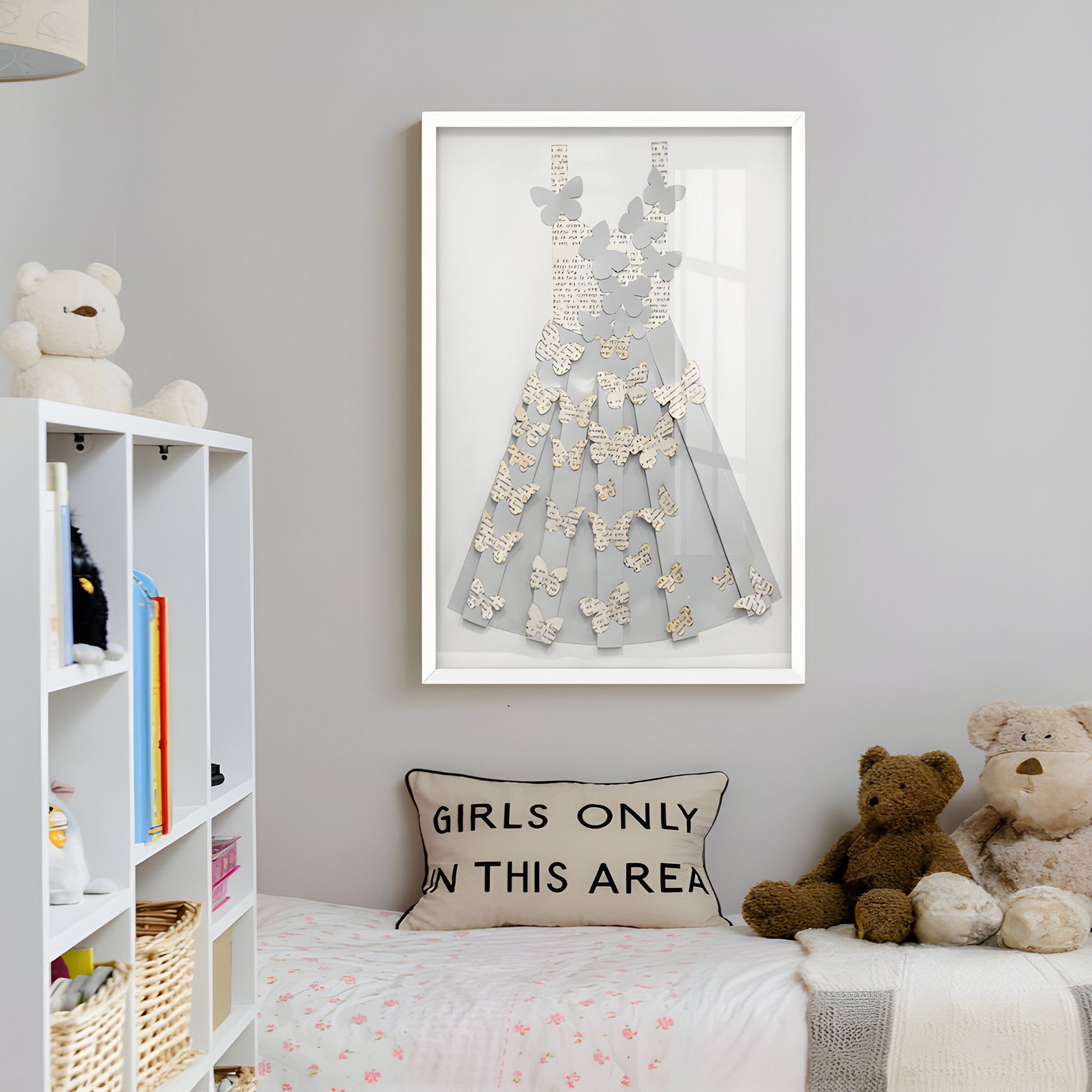 A cozy room has a bed adorned with a Girls Only In This Area pillow and teddy bears. Exquisite craftsmanship shines in the Flutter Grey & White Butterfly Dress 3D Wall Art by Giant Sculptures above. A shelf with books and a basket adds charm to the space.