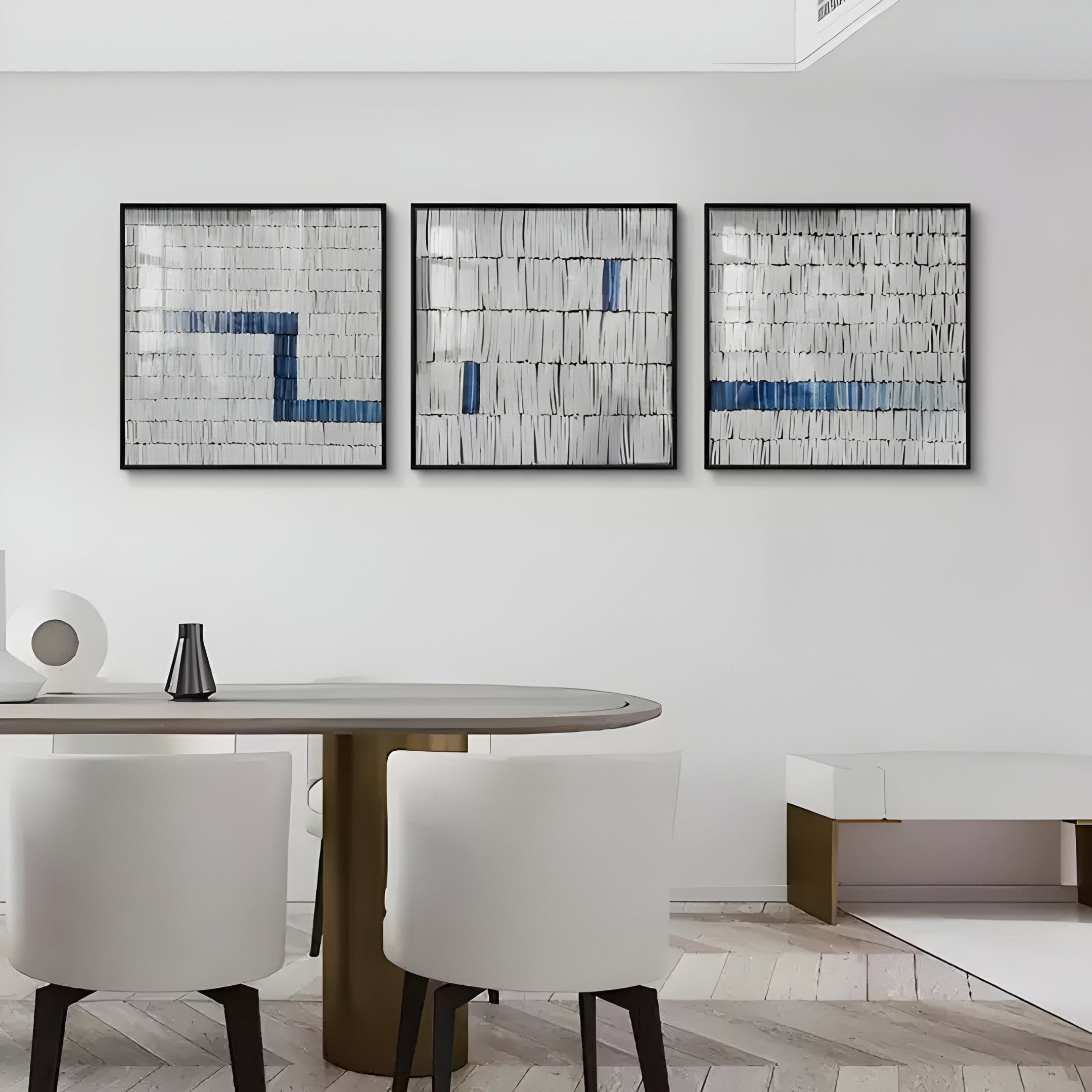 A modern dining area features a round table, white chairs, and a minimalist design. Adorning the walls are three framed Tracery I Blue & White Textured Paper Wall Art pieces by Giant Sculptures. A neutral palette complements the herringbone wood floor.