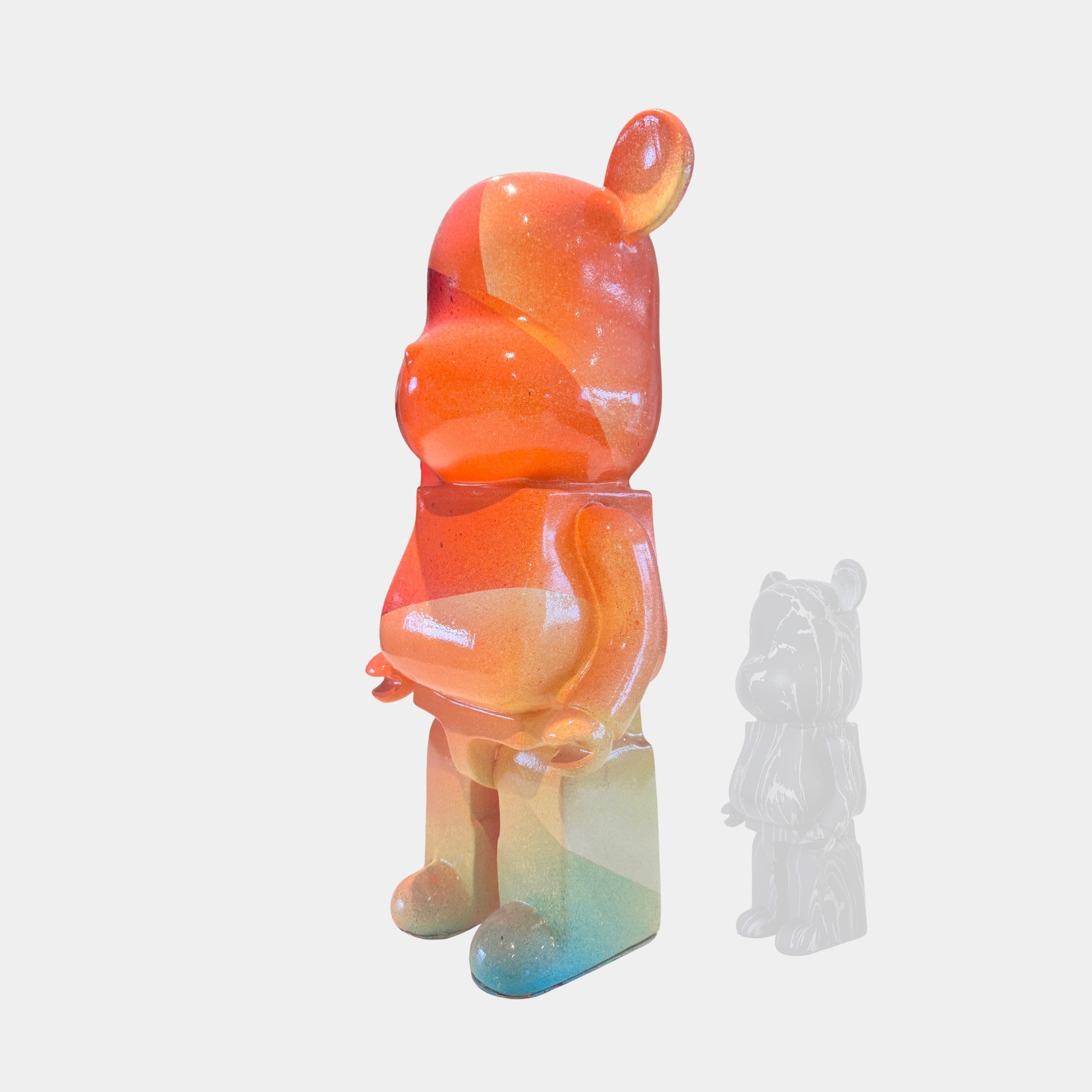 In the foreground stands the Candy Bear Sculpture - 50cm by Giant Sculptures, a bear-shaped figure with an orange, red, and blue gradient and glossy finish. Behind it sits a smaller, translucent version on a plain white background.