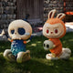 Two whimsical Giant Sculptures figurines sit on grass: the 100cm Spooky Buddies Skull Drop Labubu Sculpture in a top hat looks sad, while a smiling bunny-costumed figure holds a skull. A stone wall and wooden door suggest an indoor scene.