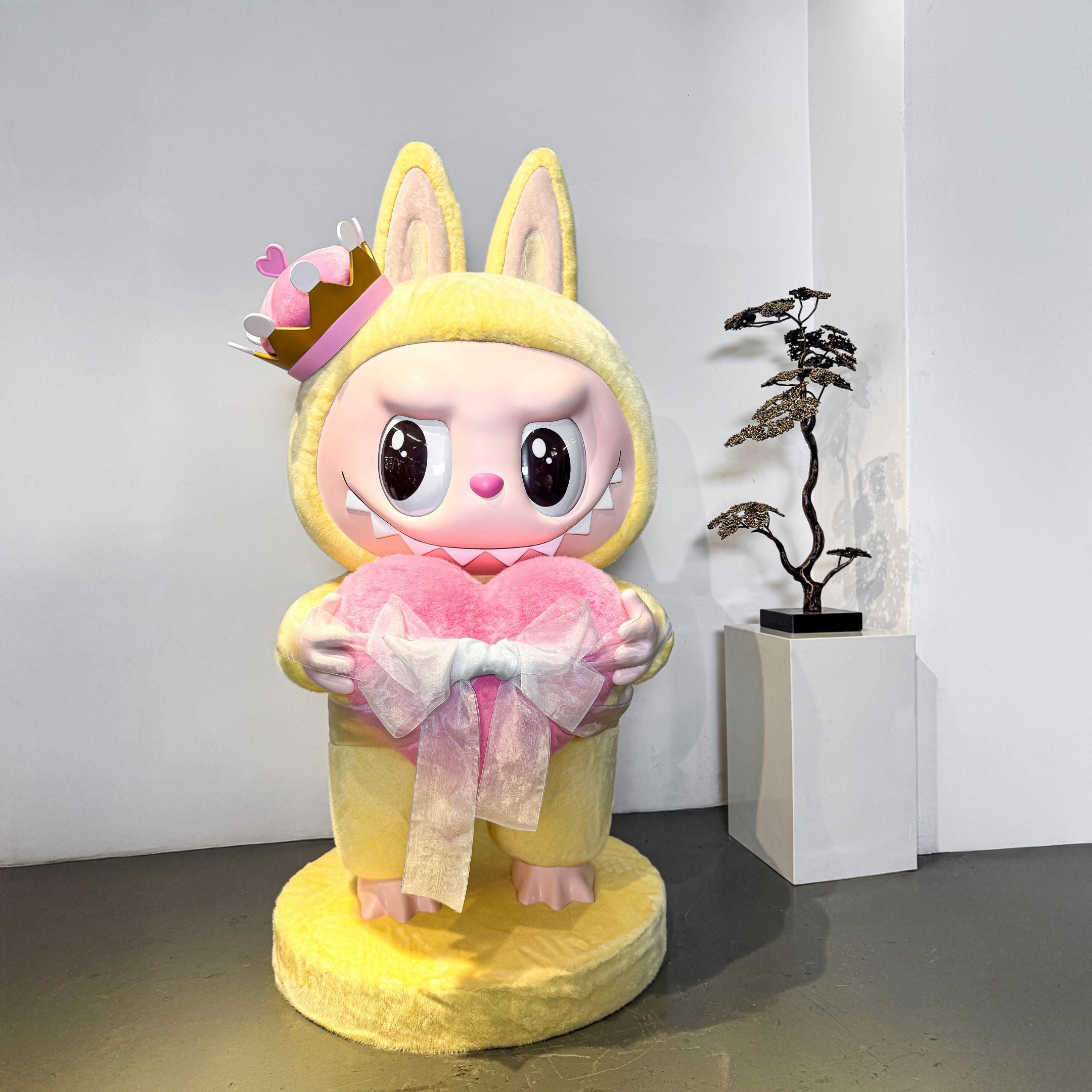 The Monster Checkmate Heart Labubu Sculpture - 100cm by Giant Sculptures features a large, cute figure with big eyes, sharp teeth, a yellow costume, a heart crown, and a pink heart with a white bow; a bonsai tree sits in the background.