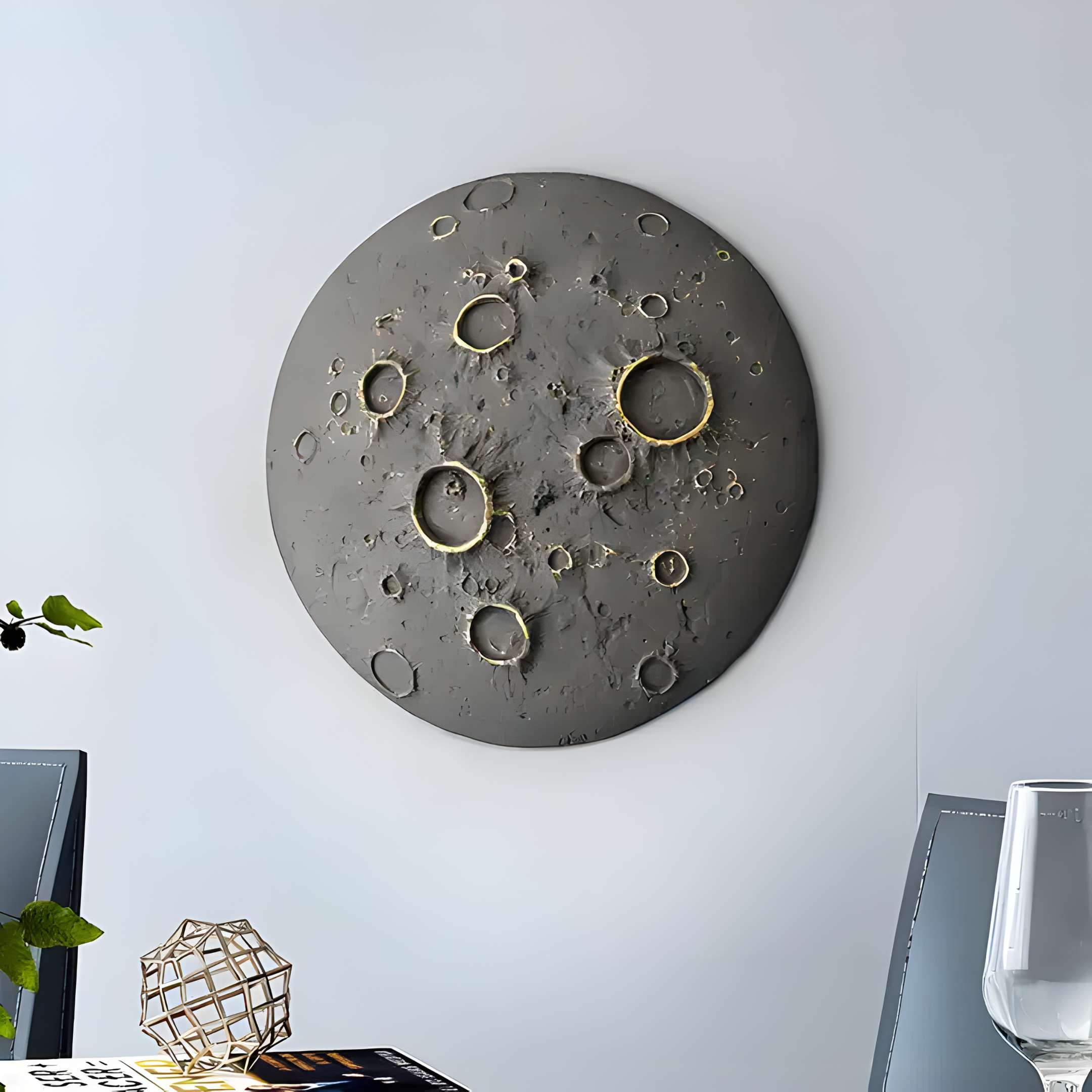 The Dark Grey 3D Lunar Impact Moon Relief Wall Art by Giant Sculptures adds cosmic décor, resembling a cratered moon. It hangs on a light gray wall above a dining table with a glass, small geometric sculpture, and an open book.