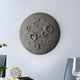 The Dark Grey 3D Lunar Impact Moon Relief Wall Art by Giant Sculptures adds cosmic décor, resembling a cratered moon. It hangs on a light gray wall above a dining table with a glass, small geometric sculpture, and an open book.