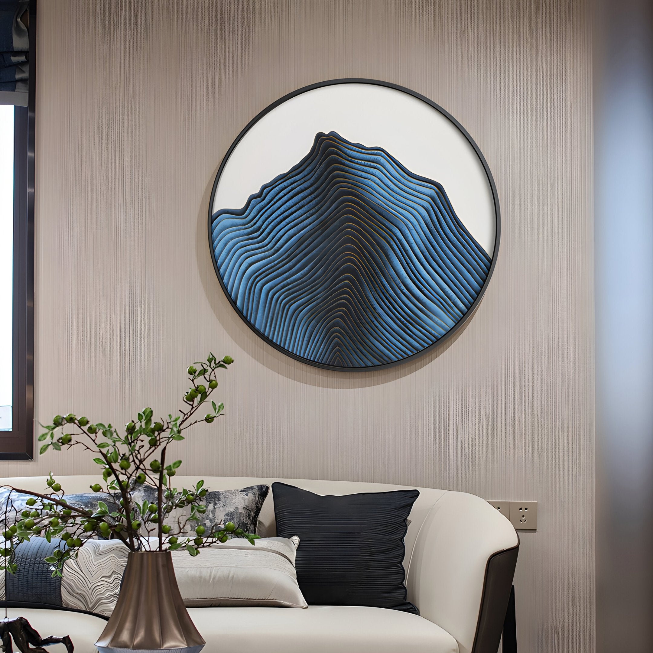 A modern living room showcases an Auric I Blue Wave Abstract Copper Wire Enamel Round Wall Art by Giant Sculptures. A white sofa with dark cushions sits below, accompanied by a small plant in a vase.