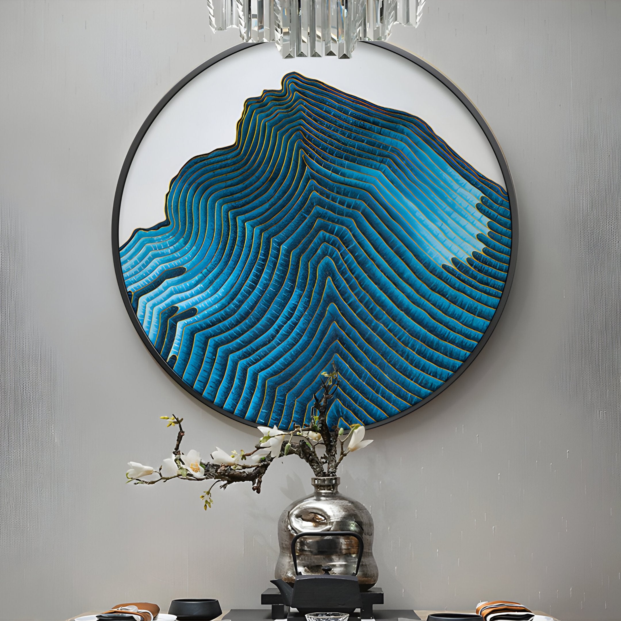 The Auric II Blue Wave Abstract Copper Wire Enamel Round Wall Art by Giant Sculptures, featuring topographic-like blue waves, adorns a gray wall. Below, a silver vase holds white blooms while a geometric chandelier enhances the arts beauty in this tranquil space.