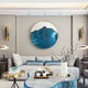 A modern living room features a sleek gray couch with blue and gold decor. The centerpiece is the Auric III Blue Wave Abstract Copper Wire Enamel Round Wall Art by Giant Sculptures. Geometric tables and a vase of white flowers are highlighted by contemporary lighting, enhancing the elegant look.