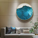 A modern living room features a beige sofa with black, blue, and white cushions. Above it hangs the Giant Sculptures Auric IV Blue Wave Abstract Copper Wire Enamel Round Wall Art with teal and blue patterns, while a plant with white flowers enhances the contemporary vibe.