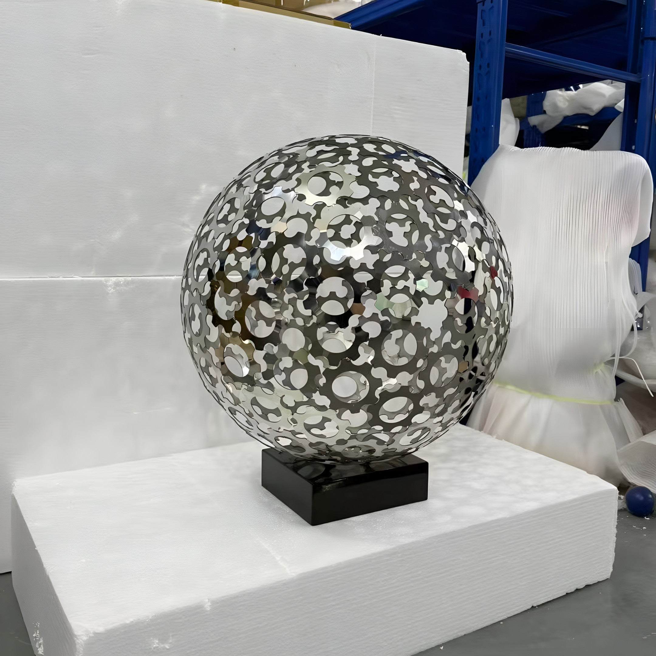 The Eclipse Silver Abstract Steel Sculpture (44/48cm) by Giant Sculptures features circular cutouts on a black base, displayed on a white foam pedestal in an industrial space with blue shelves and wrapped items in the background.