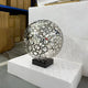 The Eclipse Silver Abstract Steel Sculpture (44/48cm) by Giant Sculptures features circular cutouts, rests on a black base atop a white block, and is displayed in a warehouse with cardboard boxes and shelves in the background.