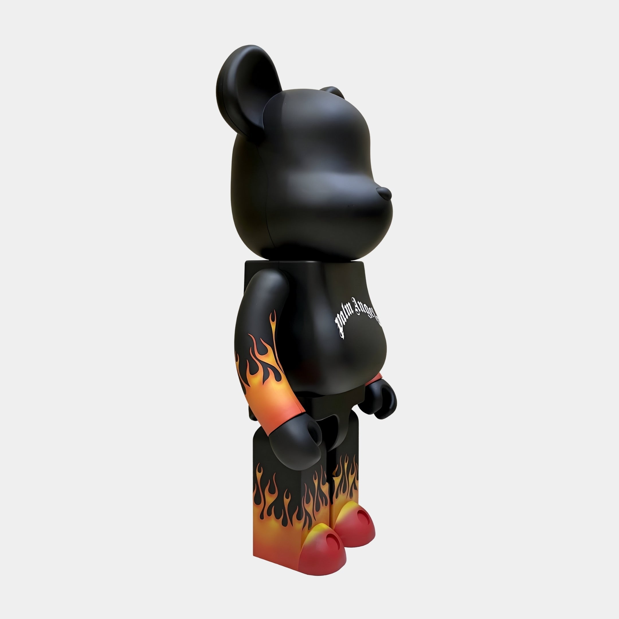 The Flame Black Bear Sculpture by Giant Sculptures is a 70cm bear-shaped collectible with a matte black finish, gradient flame details on its limbs, red feet, and a gothic Palm Angels logo on its chest, shown from the right side.