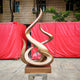 The Flame III Matt Bronze Abstract Steel Sculpture by Giant Sculptures features looping curves on a square base, displayed outdoors before two large red containers and a brick building with trees overhead.