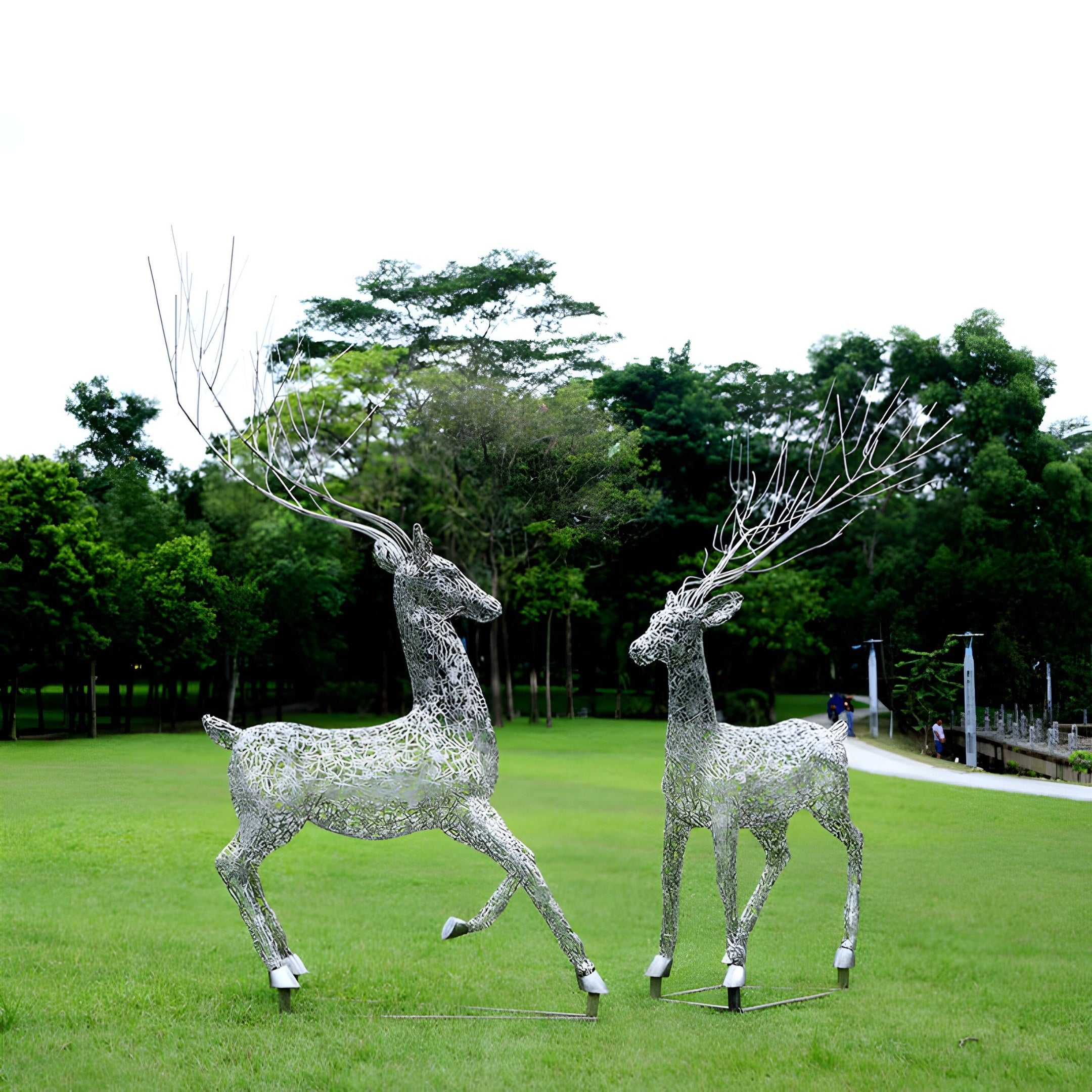 Two Giant Sculptures Arcane Stag Steel Outdoor Sculptures, each 205cm, with intricate designs and antlers reaching skyward, grace a lush lawn. Trees and a path appear in the background under clear skies, highlighting this marvel of modern art.