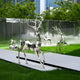 A modern garden features a collection of Noble Deer II Geometric Steel Sculptures from Giant Sculptures, crafted in stainless steel and standing prominently at 198cm tall. These artistic deer sculptures are beautifully positioned on grass with white paths, set against a backdrop of trees and buildings with mist delicately rising near a glass wall. Their varying sizes and poses bring an abstract charm to this enchanting outdoor art scene.