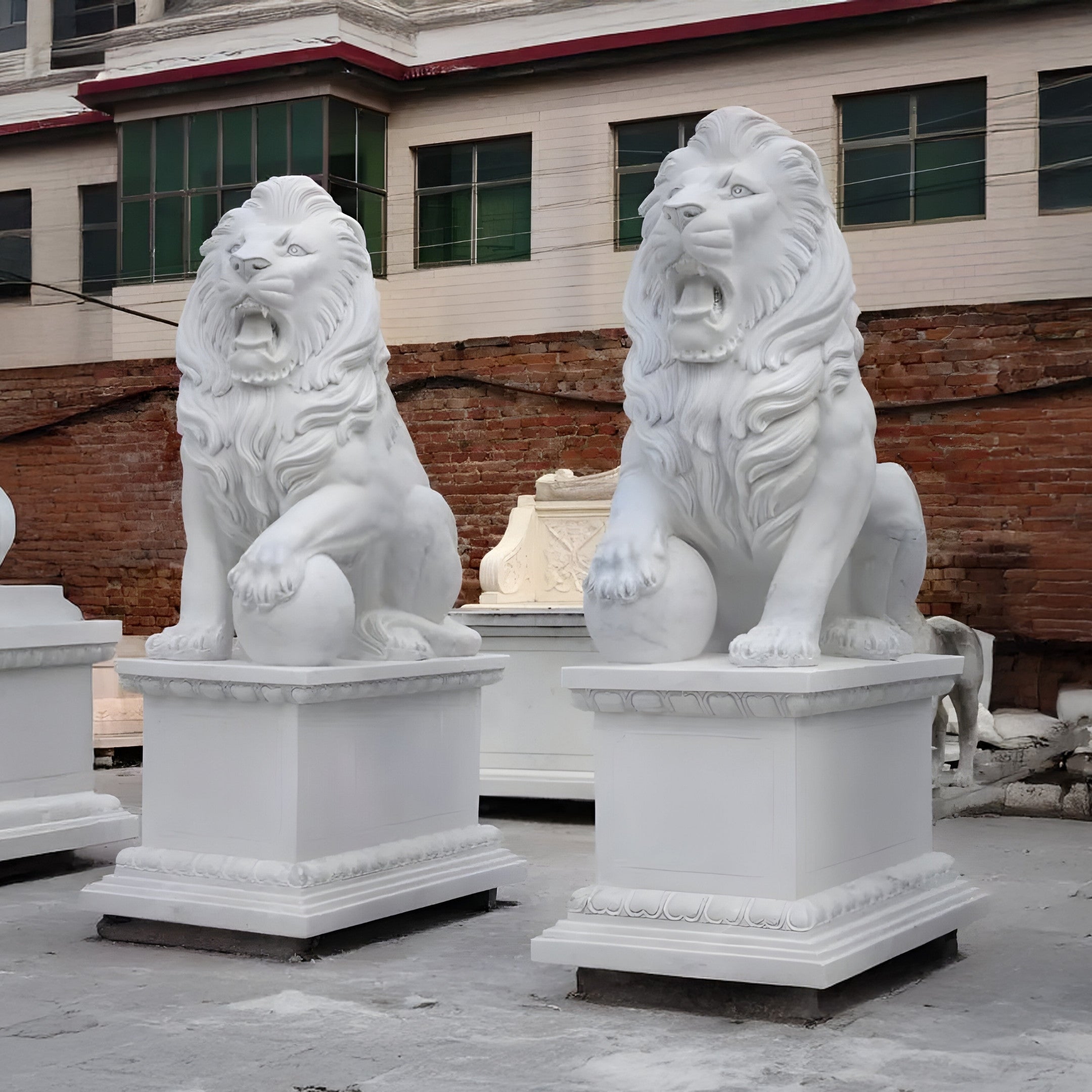 Two Giant Lion With Ball Marble Outdoor Sculptures, each 180cm tall from Giant Sculptures, majestically sit on pedestals outside the building. A brick wall and cream structure with green-tinted windows create an impressive backdrop for these customizable works of art.