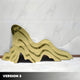 A gold, wavy abstract sculpture evokes mountain ranges with layers on a polished floor against a marble-textured wall. Intricate shadows add depth to its fluid form. The piece, labeled Version 3, is a product of Giant Sculptures titled Mountain Range Gold Abstract Steel Sculptures.