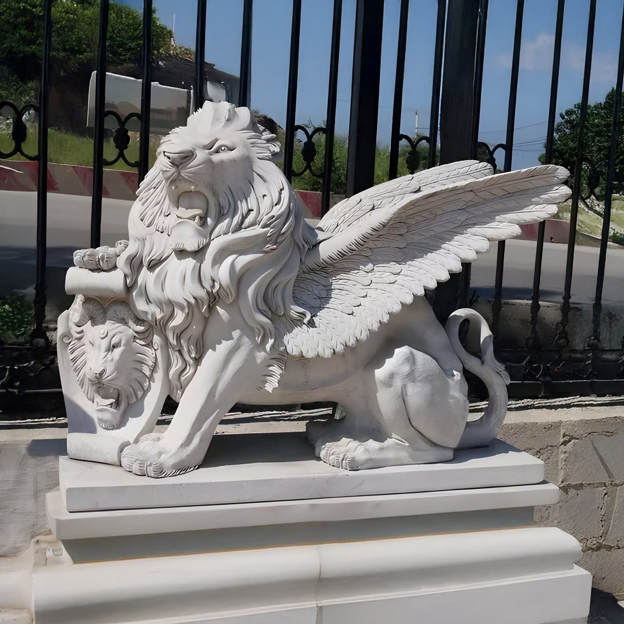 The Giant Winged Lion Marble Outdoor Sculpture (160cm) by Giant Sculptures is an impressive garden décor piece. This white marble statue depicts a winged lion with a fierce look and a scroll under its paw, standing proudly on its pedestal amid lush green bushes and wrought iron fencing.