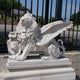 The Giant Winged Lion Marble Outdoor Sculpture (160cm) by Giant Sculptures is an impressive garden décor piece. This white marble statue depicts a winged lion with a fierce look and a scroll under its paw, standing proudly on its pedestal amid lush green bushes and wrought iron fencing.