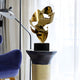 The Crest Gold Abstract Steel Sculpture by Giant Sculptures, featuring abstract gold curves, is elegantly displayed on a black pedestal. It occupies a small table beside a blue chair and tall floor lamp, glimmering as sheer white curtains allow light to fill the room.