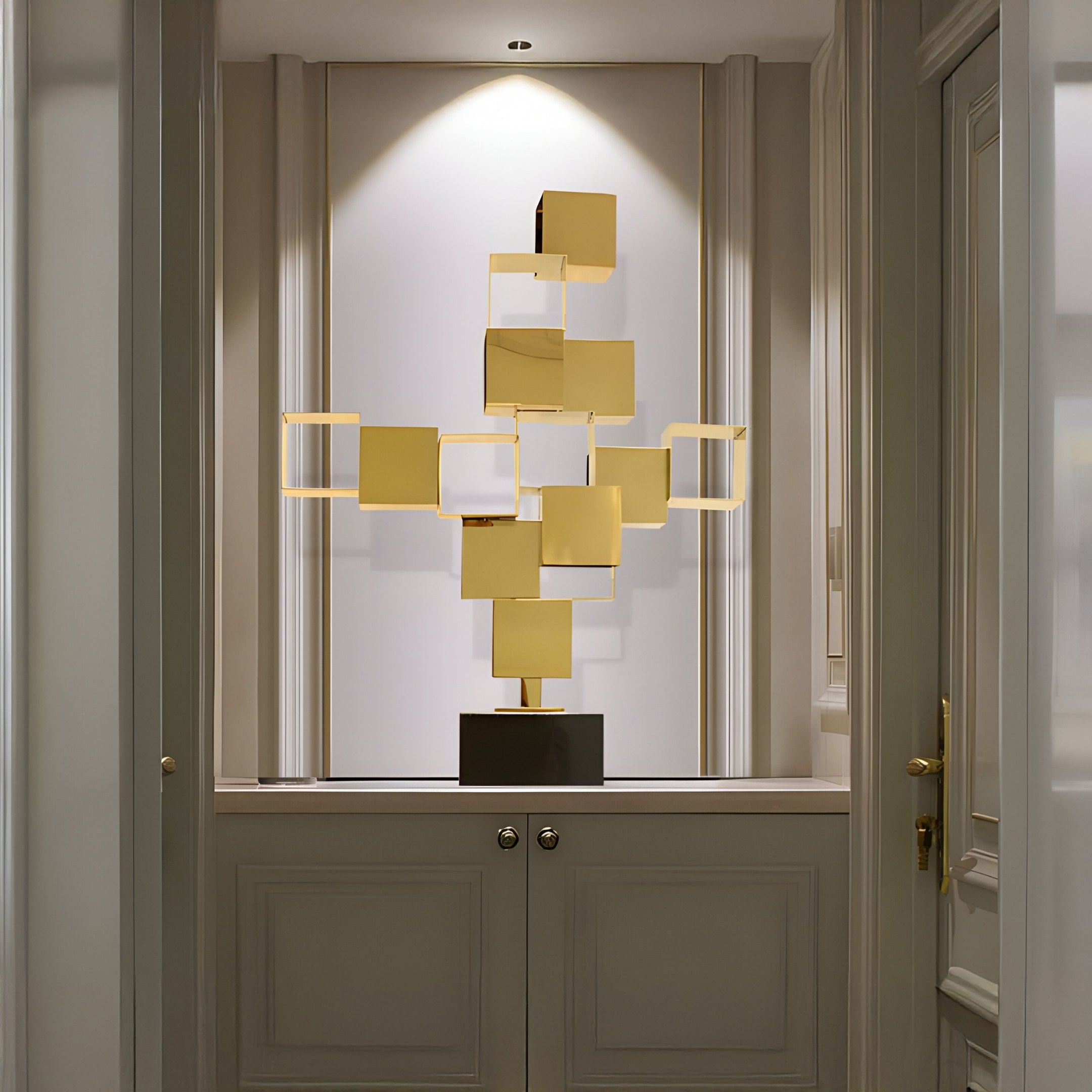 The Cuboid Gold Geometric Steel Sculpture (88cm) by Giant Sculptures features interconnected squares and rectangles on a pedestal, illuminated in a niche with soft lighting. Surrounded by neutral-toned decor, it elegantly complements both indoor and outdoor spaces.