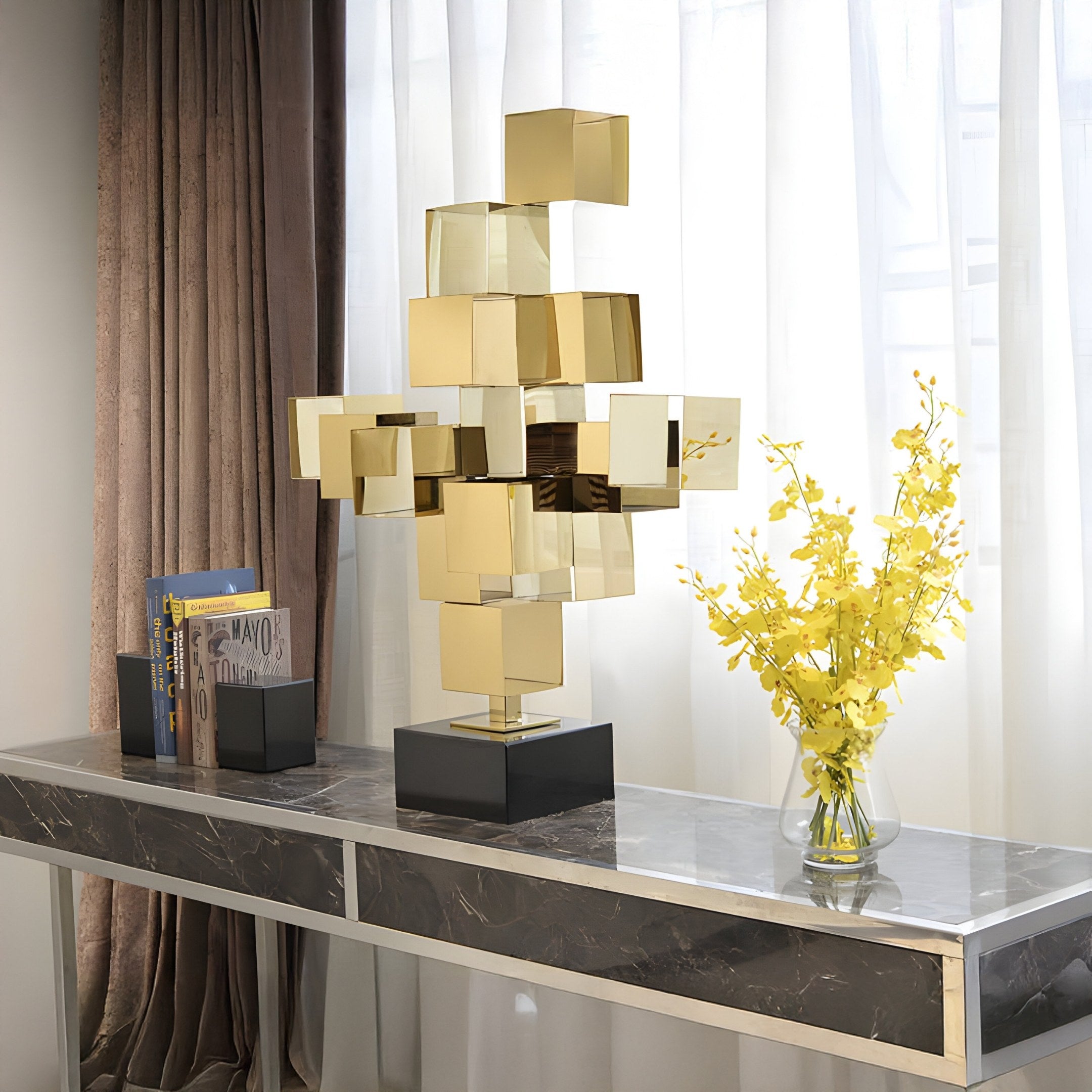 The Cuboid Gold Geometric Steel Sculpture by Giant Sculptures sits atop a marble-topped console table, accompanied by a vase of vibrant yellow flowers and book holders. Sheer curtains and brown drapes provide a seamless backdrop, blending indoor and outdoor spaces beautifully.