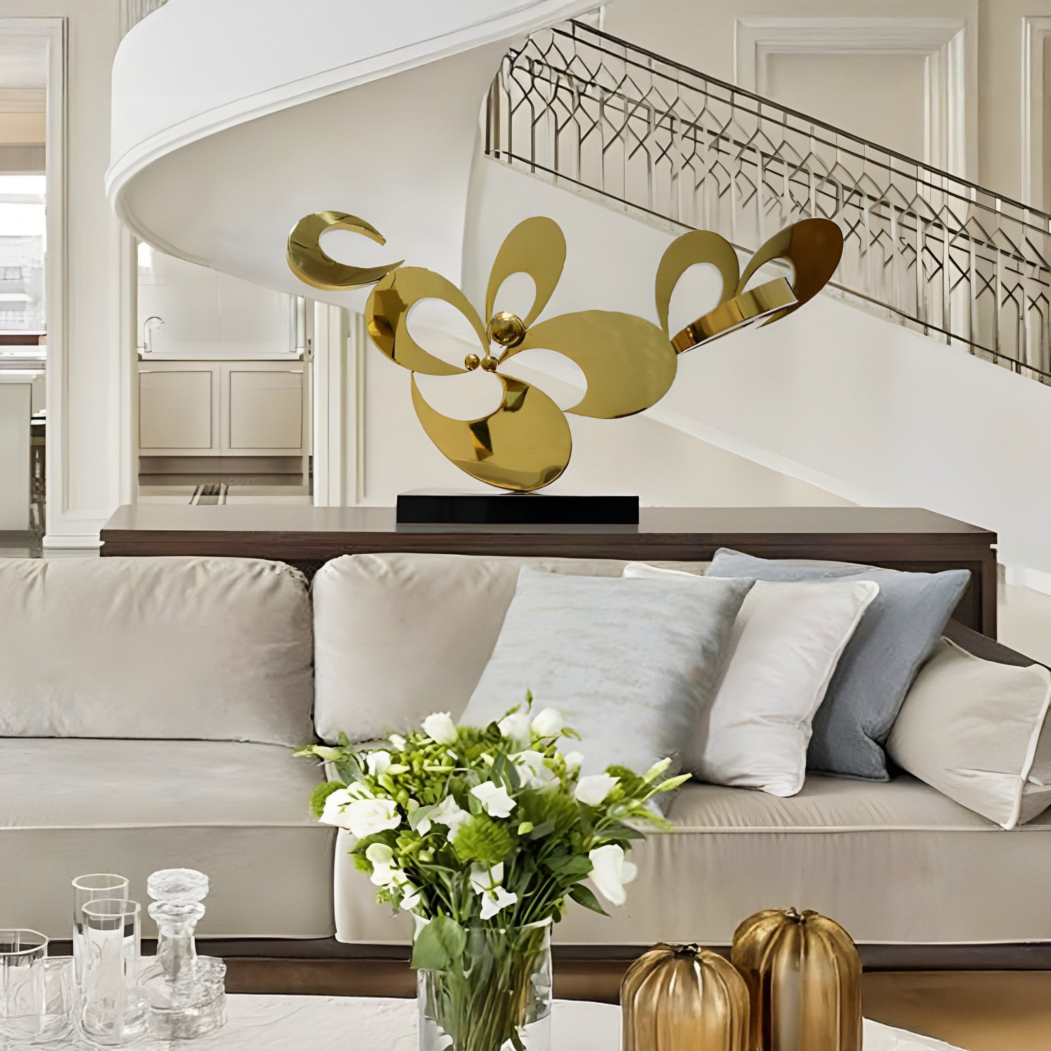 A modern living room features a beige sofa, accent pillows, and a vase of white flowers on the table. The focal point is the Cyclone Abstract Organic Steel Sculpture by Giant Sculptures displayed in front of a spiral staircase, adding elegance to the neutral-toned space.