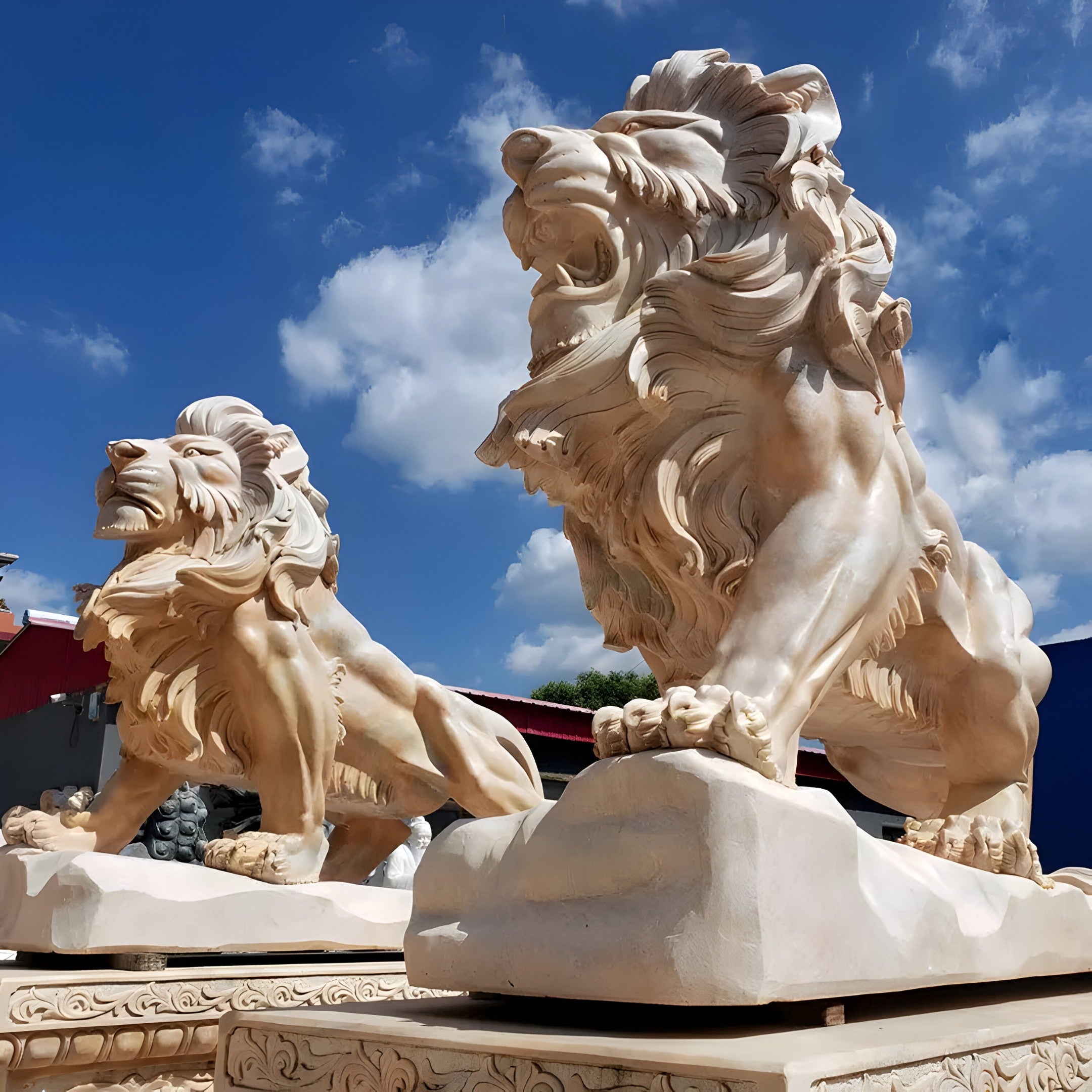 Under a bright blue sky adorned with fluffy clouds, two Giant Lion Yellow Marble Outdoor Sculptures - 240cm by Giant Sculptures stand majestically. These intricately carved and customizable pieces exhibit fierce expressions and flowing manes, positioned gracefully on decorative pedestals.