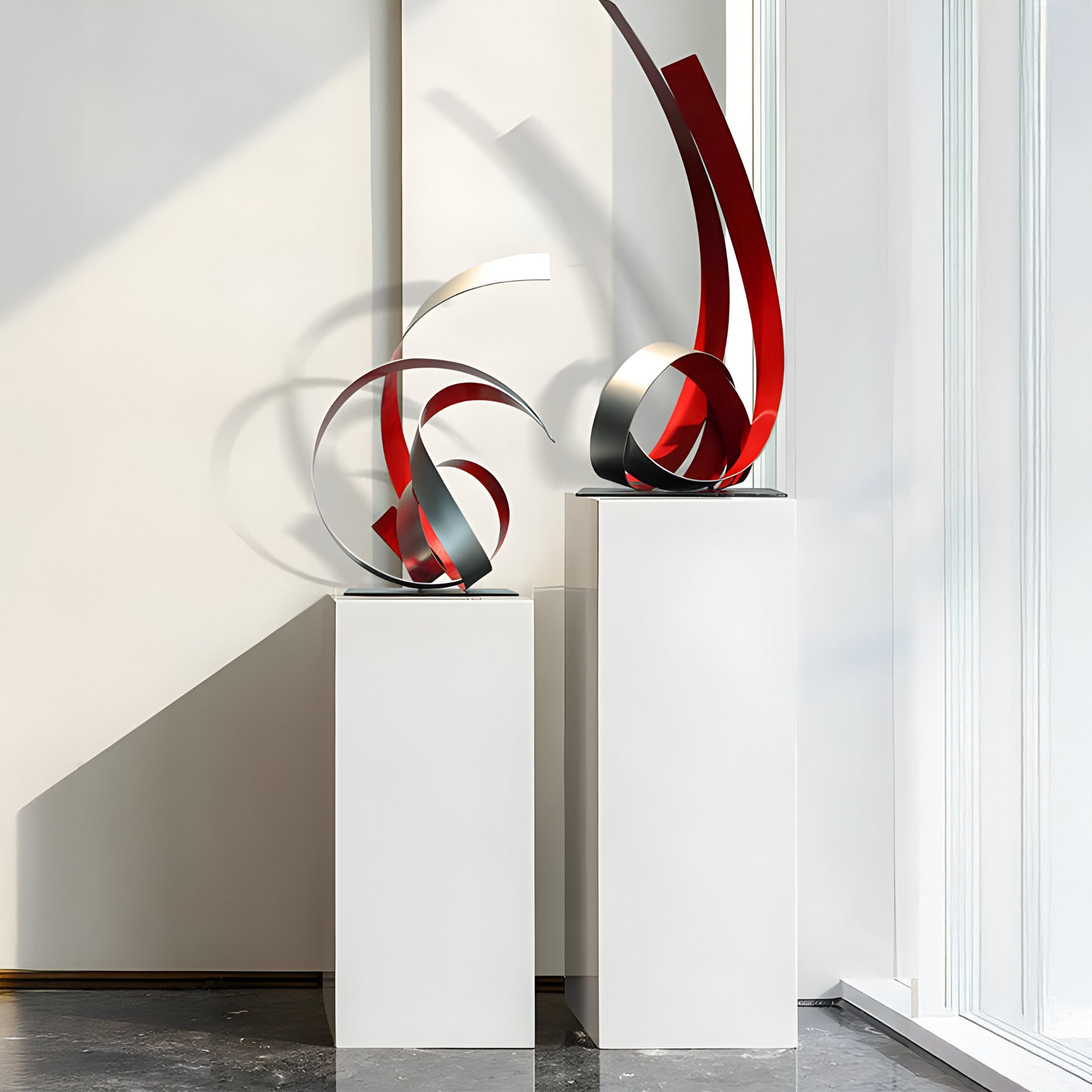 Two *Ribbon Red Abstract Steel Sculptures - 73cm* by Giant Sculptures, featuring elegant swirling ribbons of red and silver, are displayed on tall white pedestals in a sunlit gallery, casting intricate shadows on the wall behind them.