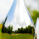 A close-up of the Raindrop Organic Steel Outdoor Sculpture by Giant Sculptures reflects a park scene with trees, grass, and a cloudy sky. The shiny surface distorts the vibrant landscape, capturing the essence of this outdoor piece beautifully.