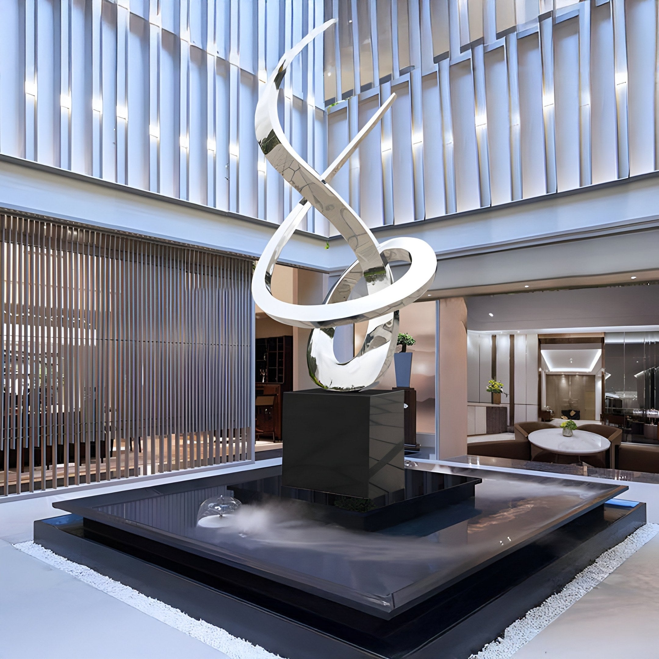 The Echo Silver Abstract Steel Outdoor Sculpture by Giant Sculptures features intertwined metallic ribbons on a black base, surrounded by water and white pebbles. This sleek 250cm structure complements minimalistic architecture with vertical slats and warm lighting.