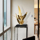 The Zephyr Gold Abstract Steel Sculpture by Giant Sculptures, standing 71cm tall, features flowing shapes on a black pedestal in a well-lit room. Its reflective surface contrasts with the neutral wall and complements the clean, contemporary decor.