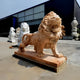 The Giant Lion II Yellow Marble Outdoor Sculpture by Giant Sculptures, standing 240cm tall with an open mouth on a wooden platform, epitomizes luxury outdoor décor. Additional lion statues enhance the grandeur near an industrial building in the background.