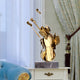 The Harmonic Gold Violin Steel Sculpture by Giant Sculptures, standing 80cm tall, sits elegantly on a marble base atop a white side table. Its positioned next to a blue sofa and framed by ornate patterned drapes with sheer white curtains behind.